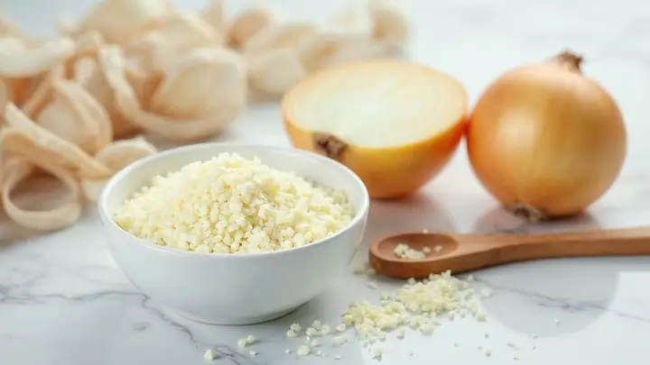 Cebola desidratada em flocos numa tigela branca ao lado de uma cebola fresca cortada ao meio sobre mármore claro